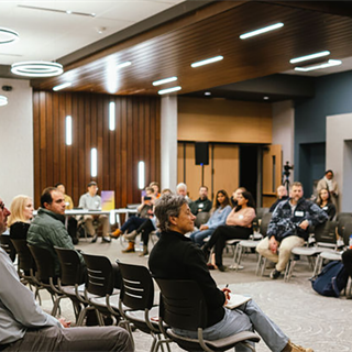 A man stands speaking before an audience divided by a wide aisle. A panel of people sit at a table a