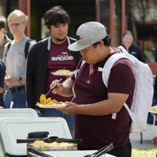 young man in red shirt, gray hat and white backpack serves himself some food from a buffet table in