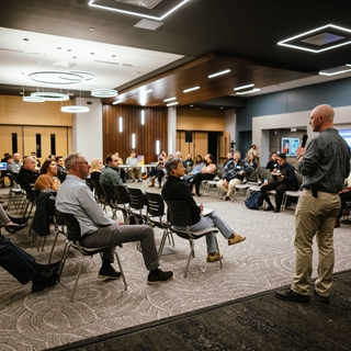 A man stands and speaks before a seated crowd in a newly remodeled room with modern lighting
