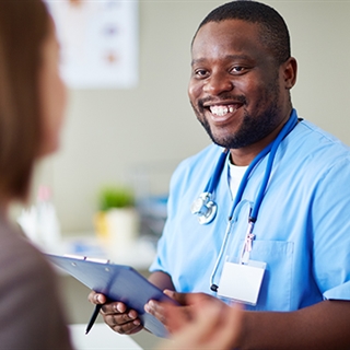 A man in blue scrubs holding a clipboard talks to a woman in the foreground facing away from the cam
